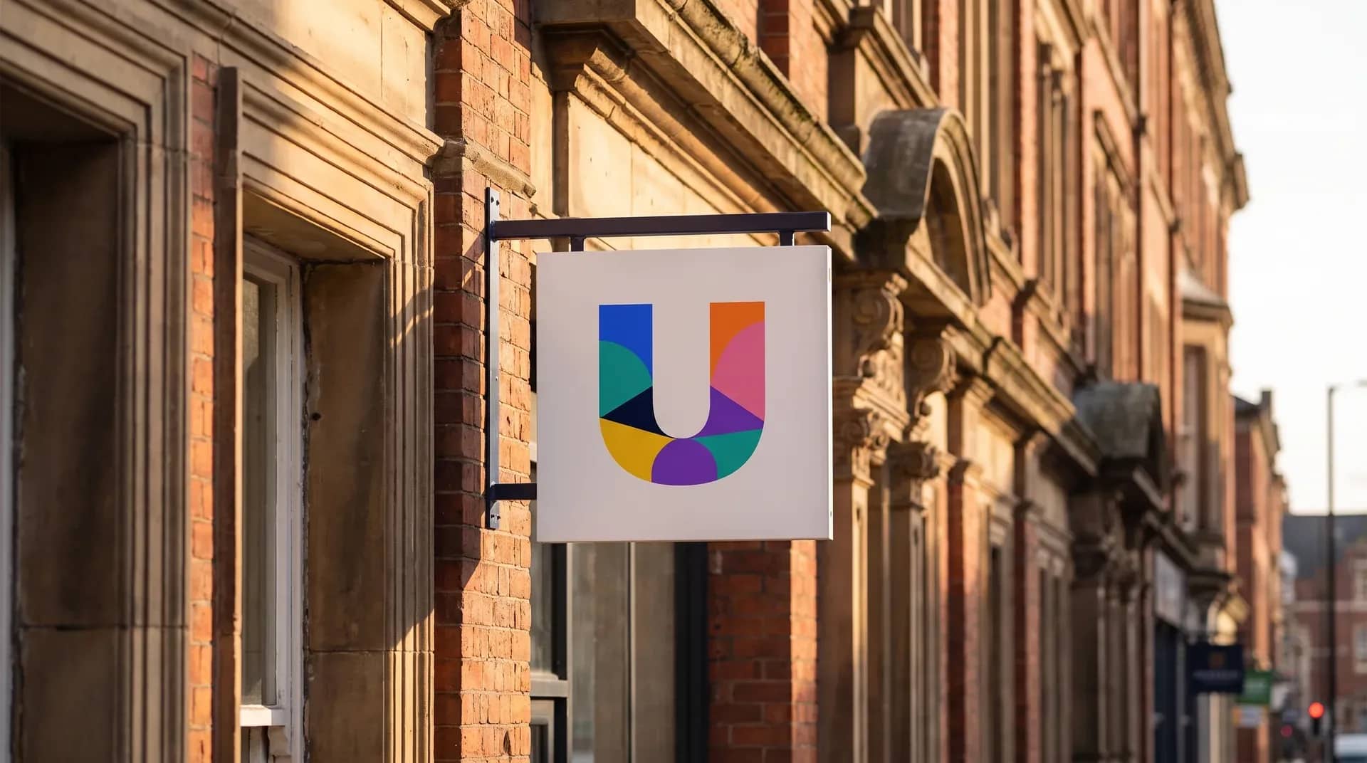 A white hanging shop sign carrying the colourful geometric UK Debt Centre 'U' mark, projecting from a red-brick Victorian high-street building in warm late-afternoon sunlight.