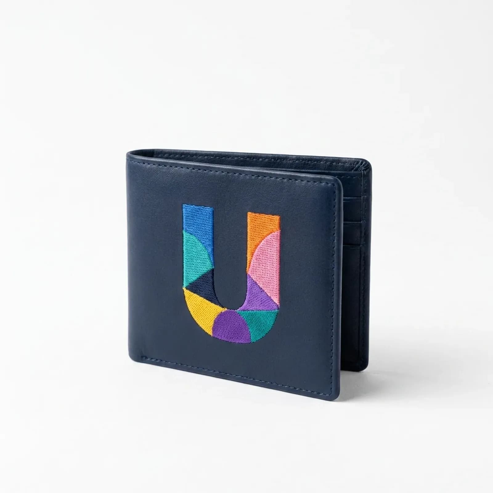 A navy leather bifold wallet standing upright on a white surface, the colourful geometric UK Debt Centre 'U' mark embroidered on its front, a soft shadow beneath it.