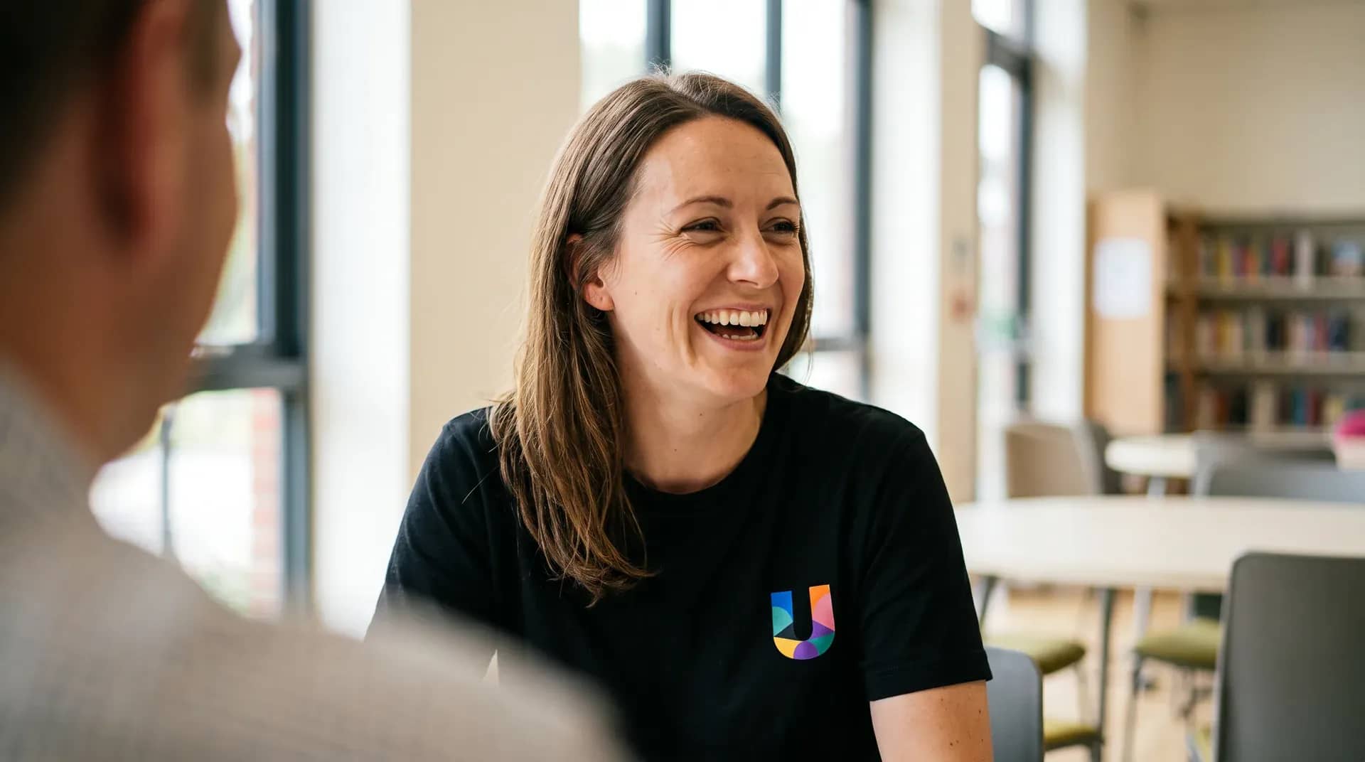 A woman in a black UK Debt Centre t-shirt laughing warmly mid-conversation with someone sitting across from her in a bright community space.