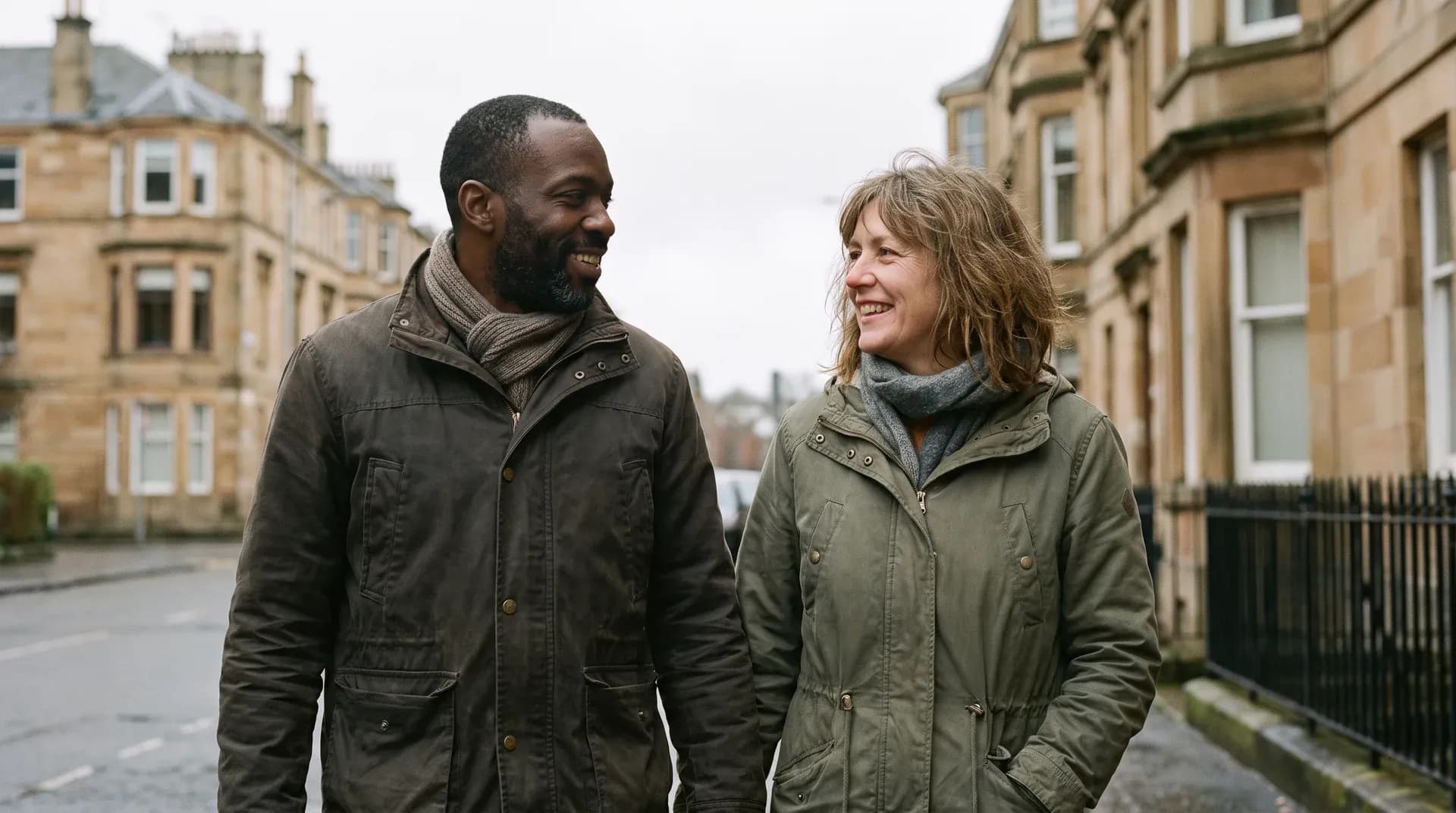 A couple in their forties walking together past sandstone tenements on a Scottish street, turning to each other mid-conversation and smiling.